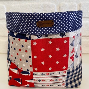 May include: A handmade fabric storage basket with a nautical theme. The basket features a patchwork design with red, white, and blue fabrics, including polka dots, stars, and sailboat patterns. The top edge is navy blue with white polka dots, and a leather tag reads "Handmade."