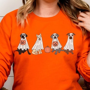 May include: An orange long-sleeved sweatshirt with a Halloween design. The graphic shows five dogs in ghost costumes, each with a pumpkin. An orange pumpkin is also included in the design. The sweatshirt is a solid orange colour.