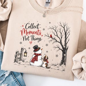 May include: A beige crewneck sweatshirt featuring a graphic print of a snowman and a dog standing near a tree and a lantern. The text reads "Collect Moments Not Things" in black and red script. The design includes snowflakes and a red bird.