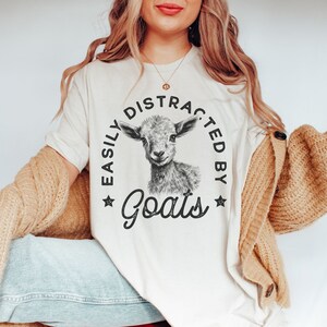 May include: A white t-shirt with a black and white illustration of a goat and the text "Easily Distracted by Goats" printed on the front.