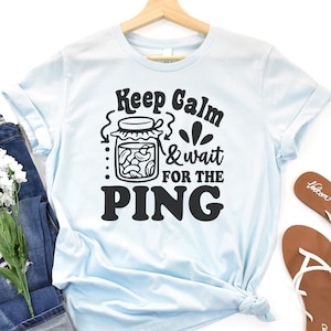 May include: Light blue t-shirt with a black graphic that says "Keep Calm & wait for the PING" with a jar of pickles illustration.