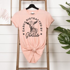 May include: A peach-colored t-shirt with a black and white illustration of a goat and the text "Easily Distracted By Goats" in a circle.