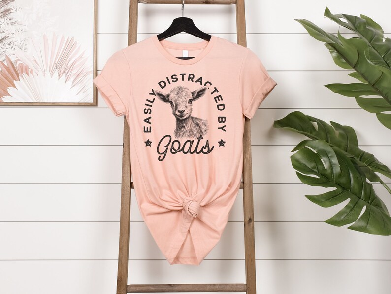 May include: A peach-colored t-shirt with a black and white illustration of a goat and the text "Easily Distracted By Goats" in a circle.