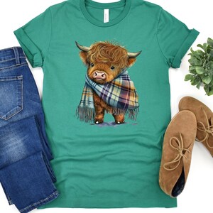 May include: A green t-shirt with a cartoon illustration of a brown and white Highland cow wearing a plaid scarf.