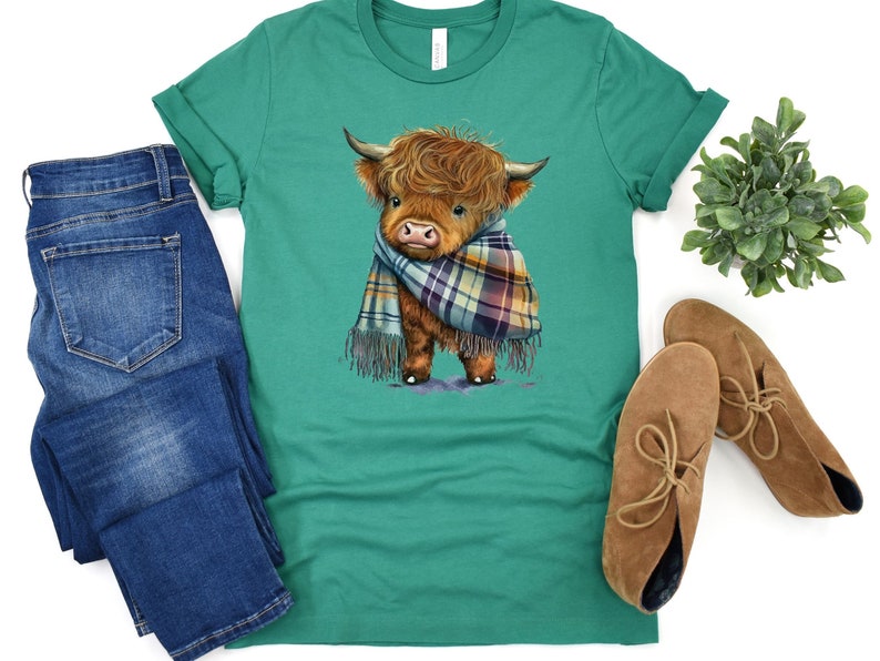 May include: A green t-shirt with a cartoon illustration of a brown and white Highland cow wearing a plaid scarf.