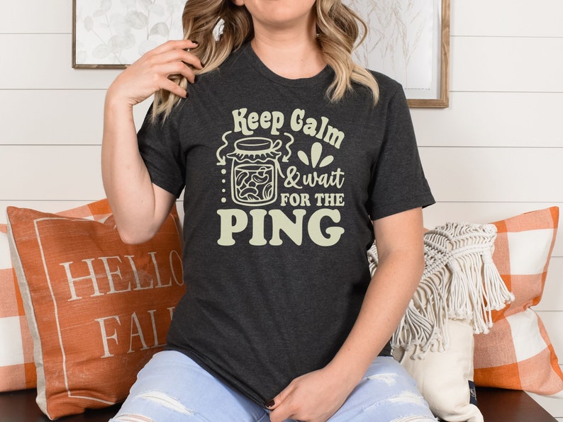 May include: A dark gray t-shirt with the text "Keep Calm & wait for the PING" in a light yellow color. The text is surrounded by a graphic of a jar of pickles.