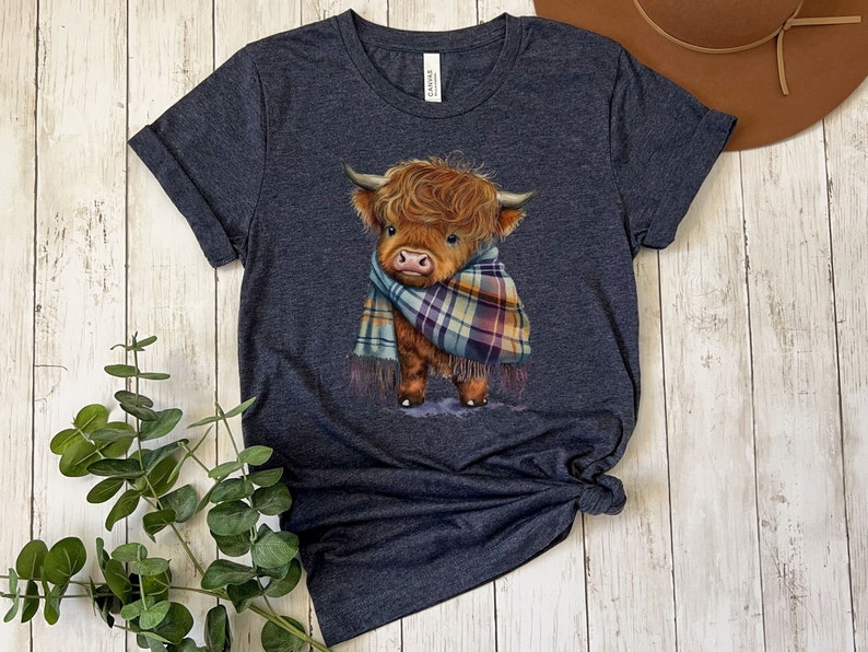 May include: A dark blue t-shirt with a cute illustration of a brown Highland cow wearing a plaid scarf. The cow is looking to the left and has a friendly expression.