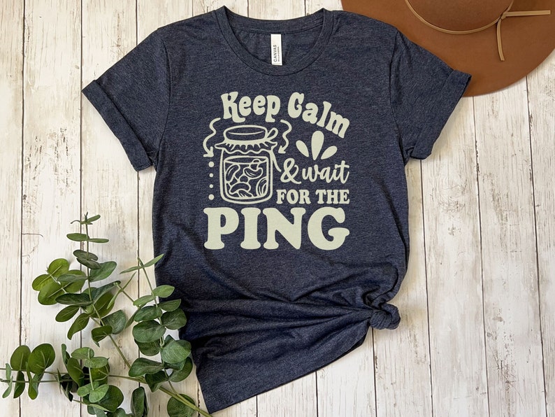 May include: A dark blue t-shirt with the text "Keep Calm & Wait For The PING" in a cream color. The text is surrounded by a graphic of a jar of pickled beans.