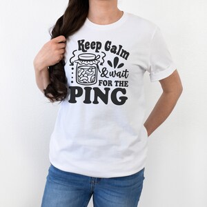 May include: White t-shirt with black text that reads "Keep Calm & Wait for the PING". The text is accompanied by a graphic of a jar filled with pasta.