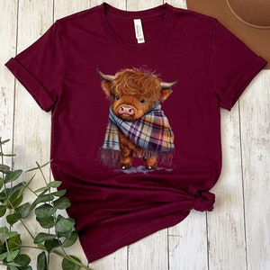 May include: A maroon t-shirt with a cute illustration of a Highland cow wearing a plaid scarf. The cow is brown and white with a fluffy mane.