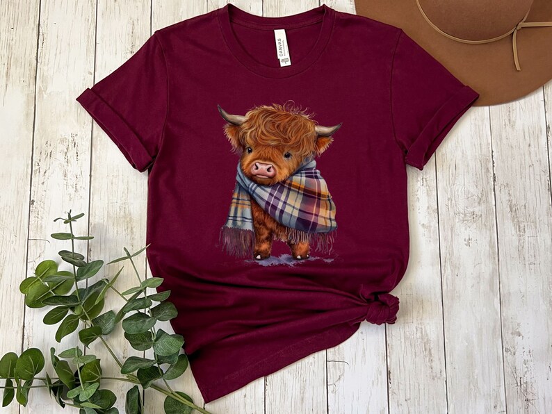 May include: A maroon t-shirt with a cute illustration of a Highland cow wearing a plaid scarf. The cow is brown and white with a fluffy mane.