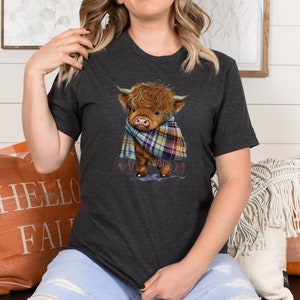 May include: A dark gray t-shirt with a cartoon illustration of a brown Highland cow wearing a plaid scarf. The cow is looking to the left and has a friendly expression.