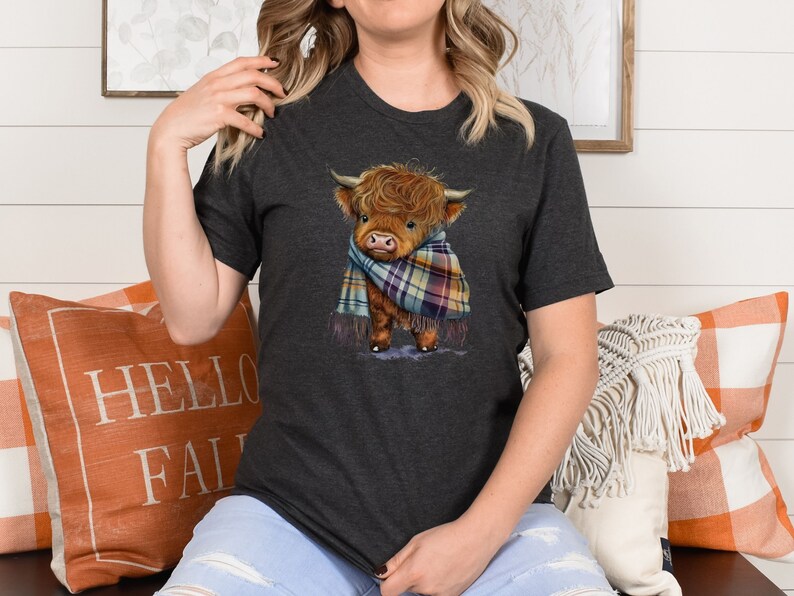 May include: A dark gray t-shirt with a cartoon illustration of a brown Highland cow wearing a plaid scarf. The cow is looking to the left and has a friendly expression.