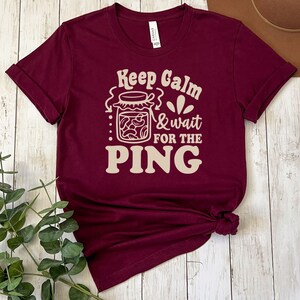 May include: A maroon t-shirt with the text "Keep Calm & Wait for the PING" in a cream color. The text is stylized with a jar of pasta and a flower graphic.