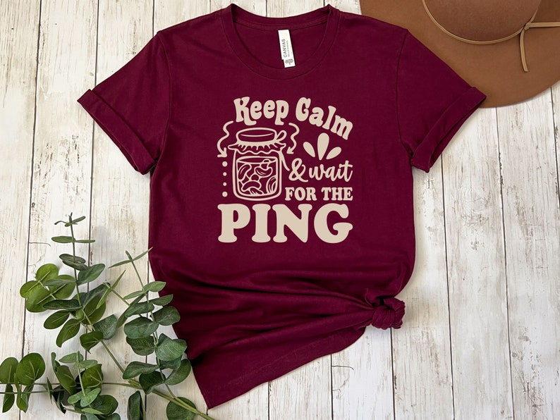 May include: A maroon t-shirt with the text "Keep Calm & Wait for the PING" in a cream color. The text is stylized with a jar of pasta and a flower graphic.