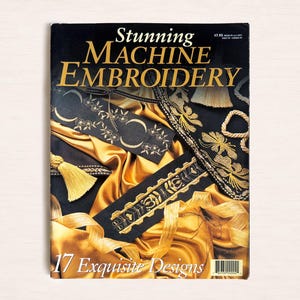May include: A magazine titled Stunning Machine Embroidery featuring 17 Exquisite Designs on the cover. The cover displays black fabric strips with gold embroidered floral and geometric patterns, accented by gold tassels and draped gold satin fabric.
