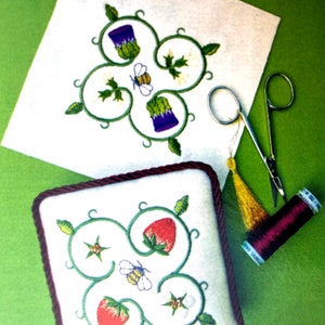 Two square pin cushions with a floral design filling the square. Each have a white background and with red and green or ourple and green motifs. A pair of small scissors and a roll of maroon cotton sit nearby. The background is green.