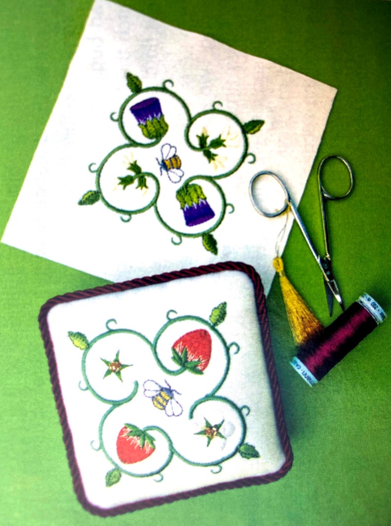 Two square pin cushions with a floral design filling the square. Each have a white background and with red and green or ourple and green motifs. A pair of small scissors and a roll of maroon cotton sit nearby. The background is green.