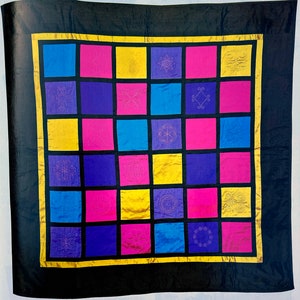 A brightly coloured square silk quilt with a black border and an internal gridded square design. Each square is bounded by a black line, making the colours stand out. The squares are blue, magenta, yellow and purple.