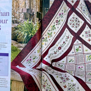 An embroidered quilt draped over a table in a garden setting. The quilt has a cream background and maroon borders, with fine maroon and green patterning. The central pattern is a diagonal grid design and there are floral motifs around the borders.