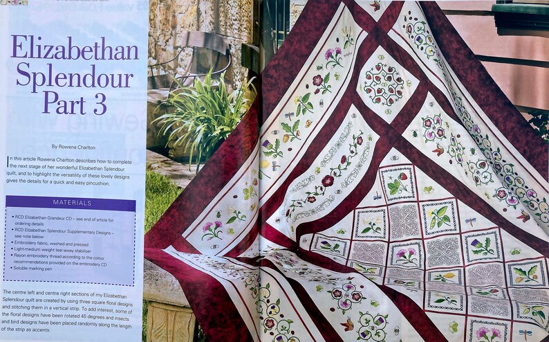 An embroidered quilt draped over a table in a garden setting. The quilt has a cream background and maroon borders, with fine maroon and green patterning. The central pattern is a diagonal grid design and there are floral motifs around the borders.