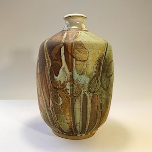 May include: A ceramic vase with a narrow neck and a rounded body featuring a multi-coloured glaze in shades of brown, tan, and muted green. The surface displays a textured, layered pattern with vertical drip-like markings and organic shapes.