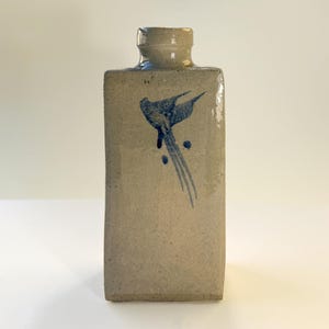 May include: A rectangular ceramic bottle with a light beige, speckled finish and a narrow neck. The front surface features a dark blue, hand-painted abstract bird motif with long, flowing tail feathers extending downwards towards the base of the vessel.