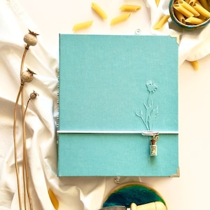 May include: A teal blue linen journal with a white ribbon closure and a small glass bottle charm. The journal has an embossed floral design and is sitting on a white surface with a small wooden knife and a teal blue ceramic plate.