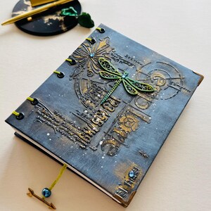 May include: A dark blue journal with a dragonfly design and gold accents. The cover features a dragonfly with green wings and a blue gem. The journal has a green ribbon bookmark and a gold arrow with a blue bead.
