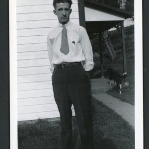 May include: Black and white photograph of a man standing outside a house. He wears a light-colored button-down shirt, dark pants, and a tie. A dog is visible in the background. The name "Carl" is written in cursive at the top of the photo.