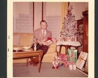 Vintage foto uit de jaren 50: een man in pak zit bij een kerstboom en houdt een ingepakt cadeau vast. Interieurdecoratie uit het midden van de 20e eeuw.