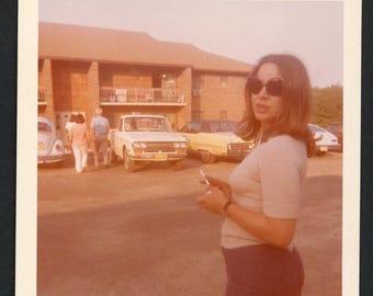 Vintage foto uit de jaren 60: vrouw met zonnebril en rokend sigaret op een parkeerplaats. Klassieke auto's.