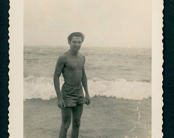 Instantánea fotográfica vintage de la década de 1940: Hombre guapo sin camisa, traje de baño, de pie en el agua de la playa.