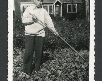 Vintage foto uit de jaren 1940: tienerjongen met hark in de hand, bladeren harkend in de achtertuin, tuinieren in de herfst.