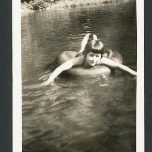 May include: A vintage black and white photograph showing a person floating on an inner tube in a lake. The person is smiling with arms outstretched. The water is calm, and trees are visible in the background.