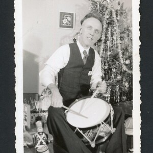 Può includere: Fotografia in bianco e nero di un uomo che suona un tamburo. Indossa un gilet, una camicia e una cravatta. Un albero di Natale decorato è sullo sfondo, con un soldatino giocattolo in primo piano.