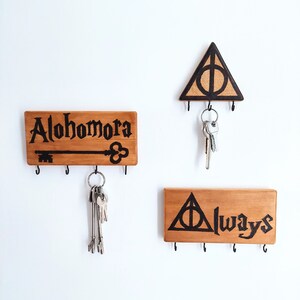 May include: Three wooden key holders with black painted Harry Potter themed designs. The top holder has a triangle with a circle inside and the text "Always". The middle holder has the text "Alohomora" and a key. The bottom holder has a triangle with a circle inside and the text "Always".