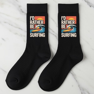 Könnte beinhalten: Paar schwarze Crew-Socken mit dem Aufdruck "I'd Rather Be Surfing". Die Grafik zeigt einen Surfer auf einem Surfbrett, der eine Welle reitet, mit einem Sonnenuntergang im Hintergrund. Die Socken sind aus weichem, bequemem Material.