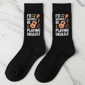 May include: Pair of black socks with the text "I'D RATHER BE PLAYING UKULELE" in white. The design includes a ukulele graphic, musical notes, and a musical staff. The socks are ribbed and likely made of a soft material.