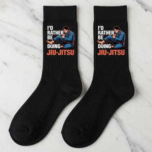Op de afbeelding: Twee zwarte sokken met de tekst "I'D RATHER BE DOING JIU-JITSU" in wit en oranje. De sokken hebben een illustratie van een persoon in een blauwe gi. De sokken zijn gemaakt van geribbelde gebreide stof.