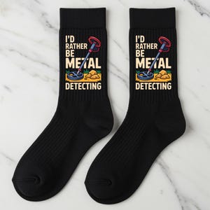May include: Pair of black socks with the text "I'd rather be metal detecting" and a graphic of a metal detector. The socks have a ribbed texture and a colorful design featuring a metal detector, grass, and rocks.