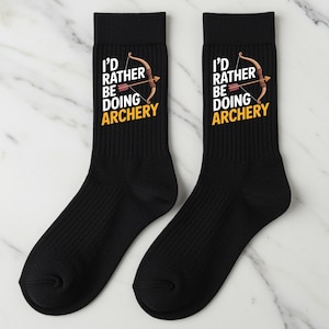 May include: Two black socks featuring the text "I'd Rather Be Doing Archery" in white and yellow, with a bow and arrow illustration. The socks are ribbed and of crew length.