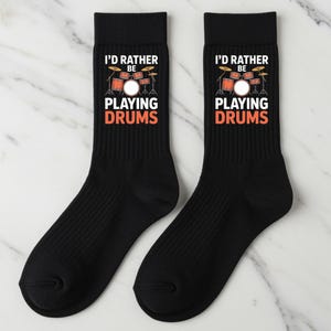 May include: Two black socks featuring the text "I'D RATHER BE PLAYING DRUMS" and a drum set graphic in orange, white, and brown. The socks have a ribbed texture and are likely made of a soft material.