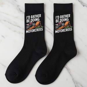 以下が含まれることがあります： 「I'D RATHER BE DOING MOTORCROSS」の文字が入った黒いクルーソックスのペア。ソックスには、木製のスロープに乗った赤と白のモトクロスのグラフィックが描かれています。ソックスは柔らかく快適な素材で作られています。
