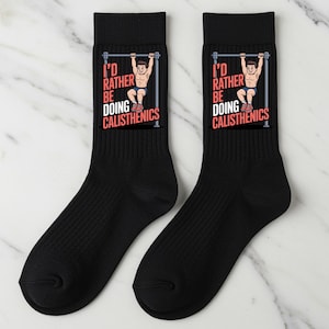 May include: Pair of black crew socks featuring a graphic of a person doing calisthenics on a pull-up bar. The text "I'd rather be doing calisthenics" is printed above the graphic in red and white. The socks are made of a soft, ribbed material.