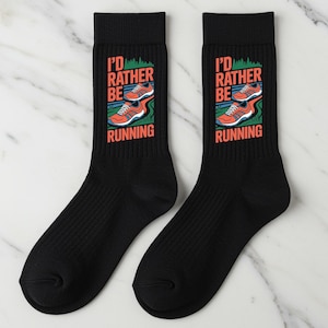 Könnte beinhalten: Paar schwarze Socken mit dem Text "I'D RATHER BE RUNNING" in Orange und Weiß. Die Socken zeigen eine Grafik von Laufschuhen in Orange, Weiß und Blau. Die Socken sind für Läufer konzipiert.