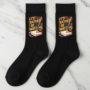 May include: Two black crew socks featuring the text "I'd Rather Be Writing" above a pencil and book illustration. The design uses yellow, red, blue, and white. A fun gift for writers.