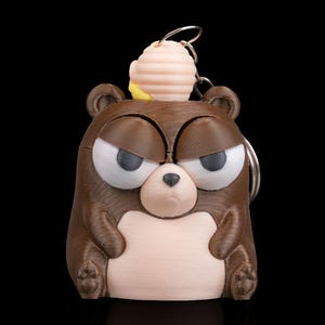 May include: A brown bear-shaped keychain with an angry expression, featuring large white eyes with dark gray eyelids and a small black nose. A light brown and yellow object sits on top of the bear's head. The keychain is made of plastic.