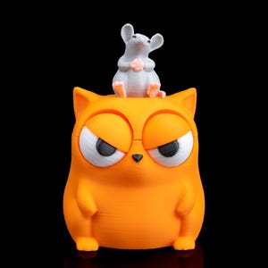May include: A 3D-printed orange cat figurine with an angry expression, featuring large white and black eyes. A small gray mouse figurine sits on top of the cat's head. The background is solid black.