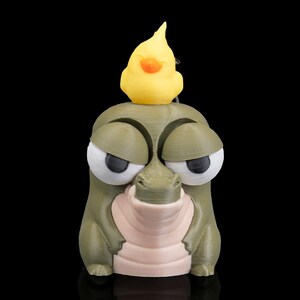 May include: A whimsical 3D-printed sculpture of a cartoonish alligator with large, expressive eyes and a yellow duck perched on its head. The alligator is primarily olive green with a light pink snout. The duck has an orange beak. The sculpture is set against a black background.
